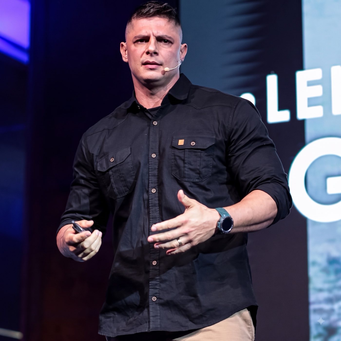 Major Ronald Cadar em palestra, no palco, gesticulando com microfone headset